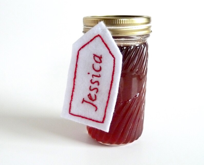 May include: A glass jar with a red liquid inside, topped with a gold lid. A white felt tag with red embroidery reads "Jessica" and is attached to the jar.