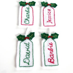 May include: Four white felt Christmas gift tags with green holly and red berries. The tags are personalized with the names Charlie, Jessica, David, and Barbie.