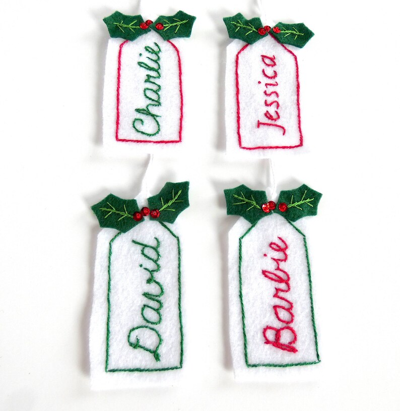 May include: Four white felt Christmas gift tags with green holly and red berries. The tags are personalized with the names Charlie, Jessica, David, and Barbie.