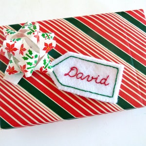 May include: A red and green striped gift box with a white bow and a white gift tag with the name "David" embroidered in red.