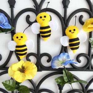 May include: Three felt bumblebees with black and yellow stripes and white wings are attached to a black metal gate with artificial flowers.