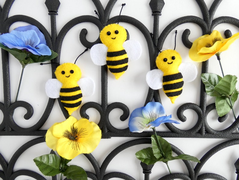 May include: Three felt bumblebees with black and yellow stripes and white wings are attached to a black metal gate with artificial flowers.