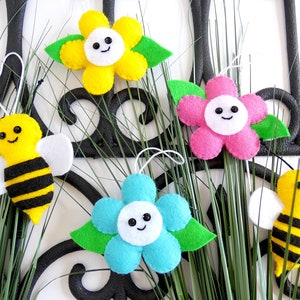 May include: Five felt ornaments, including two yellow and black bees and three flowers. The flowers are yellow, pink, and blue with white centres and smiling faces. The ornaments are hanging from green grass.