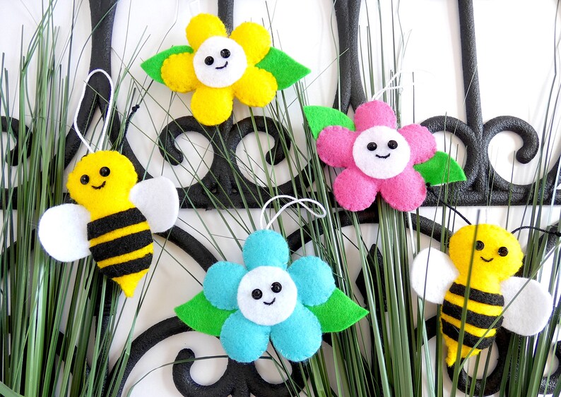 May include: Five felt ornaments, including two yellow and black bees and three flowers. The flowers are yellow, pink, and blue with white centres and smiling faces. The ornaments are hanging from green grass.
