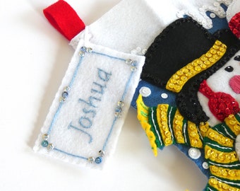 May include: A white felt Christmas stocking featuring a red hanging loop and a rectangular name tag embroidered with the name Joshua in blue thread. The stocking is decorated with a snowman wearing a black top hat and a yellow and green striped scarf.