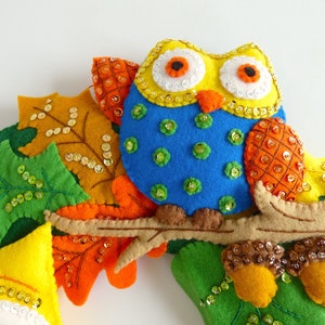 May include: A felt owl with yellow, blue, and orange accents sits on a branch surrounded by felt leaves with sequins. The owl has large, round eyes with white pupils and orange irises.