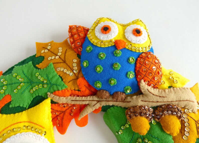 May include: A felt owl with yellow, blue, and orange accents sits on a branch surrounded by felt leaves with sequins. The owl has large, round eyes with white pupils and orange irises.