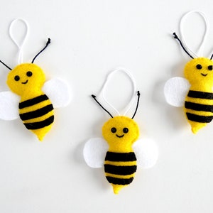 May include: Three yellow felt bee ornaments with black stripes and white felt wings. Each bee has a smiling face with black eyes and a black mouth.