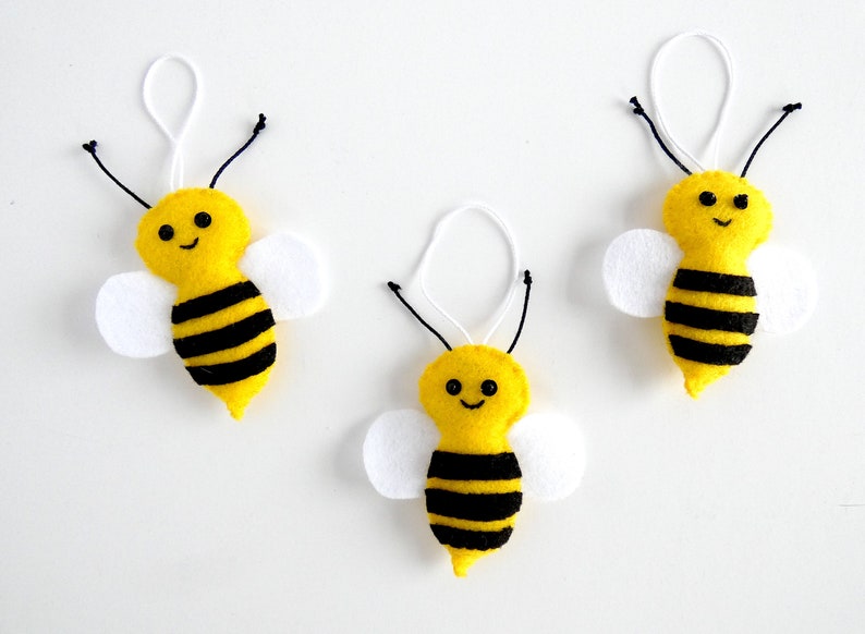 May include: Three yellow felt bee ornaments with black stripes and white felt wings. Each bee has a smiling face with black eyes and a black mouth.