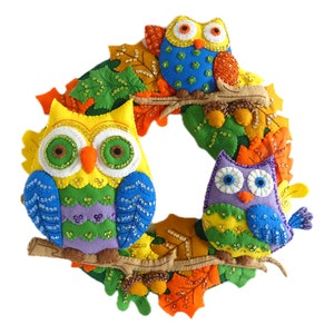 May include: A colorful felt wreath with three owls decorated with sequins. The owls are yellow, green, purple, and blue. The wreath is decorated with felt leaves in shades of orange, green, and yellow.