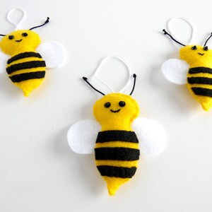 May include: Three yellow felt bee ornaments with black stripes and white wings. Each bee has a black embroidered smile and black button eyes.