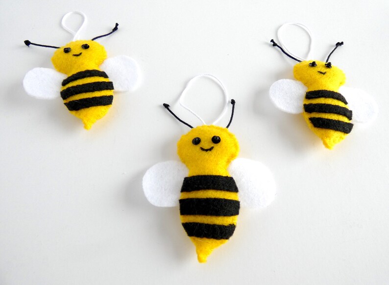 May include: Three yellow felt bee ornaments with black stripes and white wings. Each bee has a black embroidered smile and black button eyes.