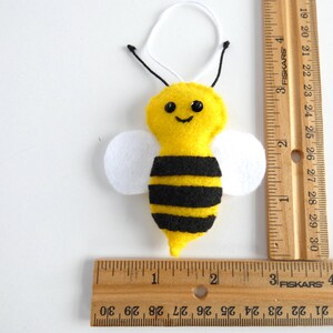 May include: A yellow felt bee ornament with black stripes, white felt wings, and black bead eyes. The bee is approximately 5 cm tall and is hanging from a white string.