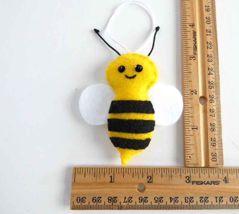May include: A yellow felt bee ornament with black stripes, white felt wings, and black bead eyes. The bee is approximately 5 cm tall and is hanging from a white string.