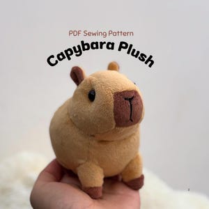 Puede incluir: Un peluche de capibara color canela con un hocico marrón oscuro y orejas pequeñas. El texto sobre el peluche dice PDF Sewing Pattern Capybara Plush. El juguete tiene ojos negros y patas marrón oscuro, con una forma corporal suave y redondeada.