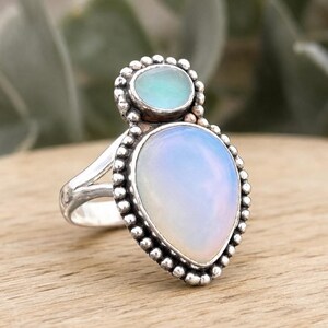 May include: A silver ring featuring two iridescent, milky white stones set in a beaded metal frame. The larger stone is pear-shaped, while the smaller stone above it is oval. The band is split, and the metal has a polished, textured finish.
