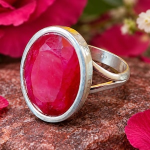 May include: A silver ring featuring a large, oval-shaped, faceted ruby gemstone set in a smooth bezel. The band is made of polished silver and includes a visible 925 hallmark stamp on the inner side of the shank, indicating sterling silver composition.