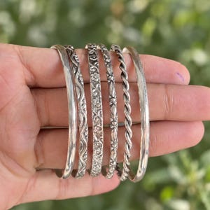 May include: A set of five silver-toned metal bangles featuring various designs, including a smooth band, a wavy pattern, a floral motif, a slender textured band, and a twisted rope-style design, all displayed together in a stacked arrangement.