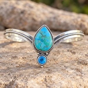 May include: A silver cuff bracelet featuring a central teardrop-shaped turquoise stone with natural veining, positioned above a smaller, round, faceted blue gemstone. The metal band has a twisted rope texture along the edges and a V-shaped centre.