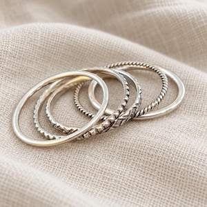 May include: A set of five silver-toned stacking rings featuring varied textures. The collection includes a smooth plain band, a twisted rope-style band, and three bands with distinct geometric, notched, and engraved patterns in a metallic finish.
