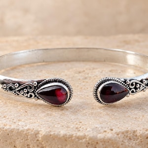 Puede incluir: Una pulsera de brazalete de plata con dos piedras de granate rojo oscuro en forma de lágrima en los extremos abiertos. La banda de metal está decorada con intrincados patrones de cuerda trenzada y pequeños detalles de cuentas de plata.