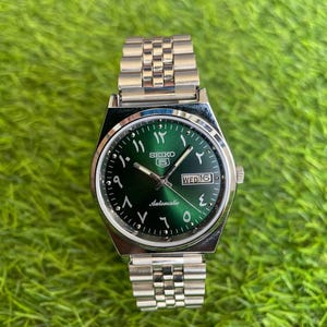 May include: A silver-tone stainless steel Seiko 5 automatic wristwatch featuring a sunburst emerald green dial with Eastern Arabic numerals, silver hands, and a day-date window at the three o'clock position, attached to a metal link bracelet.