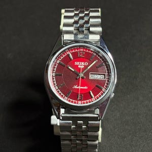 May include: A stainless steel Seiko 5 automatic wristwatch featuring a vibrant red sunburst dial with silver-tone hour markers, a day-date window at the three o'clock position, and a metallic link bracelet with a polished and brushed finish.