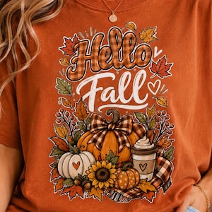 Könnte beinhalten: Ein kurzärmeliges T-Shirt in gebranntem Orange mit einem Grafikdruck und dem Text Hello Fall. Das Design zeigt Kürbisse, eine Sonnenblume, einen Kaffeebecher und Herbstblätter in den Farben Orange, Gelb, Braun und Weiß mit Karomustern.