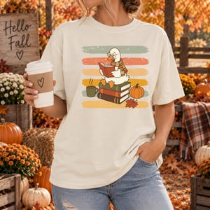 Puede incluir: Una camiseta de color crema con el gráfico de un ganso blanco con gafas y una bufanda de cuadros, leyendo un libro sobre una pila de tres libros. Junto a los libros hay una taza de café, una calabaza y una hoja de arce sobre franjas horizontales.