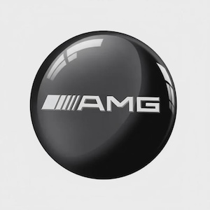 Puede incluir: Un emblema circular de color negro brillante que presenta el logotipo AMG de color plateado centrado horizontalmente. La superficie tiene un acabado liso y reflectante con un reflejo de luz distintivo en la curva superior, resaltando su forma redondeada.