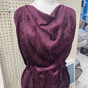 Può includere: Un top senza maniche di colore viola prugna intenso con scollo ad anello e un motivo paisley testurizzato su tutto il tessuto. Il capo è esposto su un manichino e presenta una silhouette drappeggiata con tessuto arricciato sulle spalle.