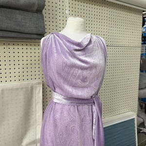 Può includere: Un abito senza maniche color lavanda con un sottile motivo cachemire e una consistenza vellutata. Il capo presenta uno scollo drappeggiato e una cintura in tessuto abbinata in vita, creando una silhouette fluida su un manichino.