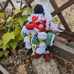 May include: A crocheted amigurumi figure of a tengu with a red face, long nose, and white hair. It wears a small black cap, a green kimono-style top with white pom-poms, and brown sandals. The figure has black eyes and a pink cheek detail.