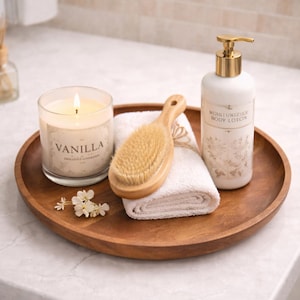 Op de afbeelding: Een rond houten dienblad met een brandende kaars in een glazen pot met het label Vanilla, een houten lichaamsborstel met natuurlijke haren, een opgevouwen wit washandje en een witte pompflacon met het label Moisturizing Body Lotion.