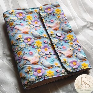 Peut inclure: Une couverture de carnet en tissu avec un fond bleu clair et un motif répétitif d'oiseaux roses et gris perchés sur des branches parmi des fleurs jaunes, violettes et roses. La couverture comporte un bouton-pression argenté.