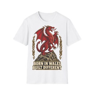 Op de afbeelding: Een wit t-shirt met korte mouwen en een afbeelding van een rode draak die op een rots staat. Het ontwerp bevat gouden Keltische knoopwerkranden aan de zijkanten en de tekst BORN IN WALES BUILT DIFFERENT in vetgedrukte letters.