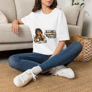 May include: A white short-sleeved t-shirt featuring a graphic of a person with messy hair sitting in bed holding a coffee mug. The text reads: I DON'T LIKE MORNING PEOPLE OR MORNINGS OR PEOPLE. The shirt is paired with blue denim jeans and white sneakers.