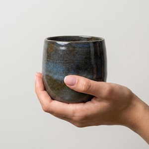 Könnte beinhalten: Ein Keramikbecher mit abgerundeter, henkelloser Form und einer gesprenkelten Glasur in tiefen Tönen von Dunkelblau, Anthrazit und erdigem Braun. Die Oberfläche hat ein glattes, glänzendes Finish mit subtilen Farbvariationen auf der Außenseite.