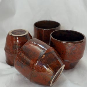 May include: A set of four ceramic cups with a glossy, deep reddish-brown glaze featuring dark, mottled textures. The cups have a slightly tapered, cylindrical shape with a smooth rim and a visible light-colored base on the bottom of the vessels.