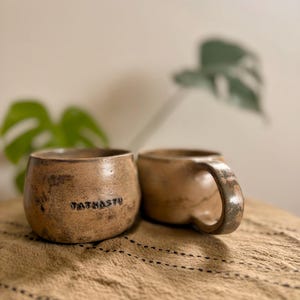 May include: Two handmade ceramic mugs with a rustic, speckled tan and brown finish. One mug features the word "TATHASTU" stamped clearly into the side. Both mugs have a rounded, organic shape with a thick, sturdy handle on the right side.