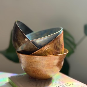 Pode incluir: Uma pilha de três tigelas de cerâmica com acabamentos variados. A tigela inferior é de um castanho-âmbar quente e salpicado. A tigela do meio apresenta uma superfície texturizada castanho-dourada. A superior tem tons metálicos escuros.