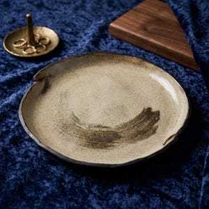 Op de afbeelding: Een ronde, beige keramische plaat met een onregelmatige, donkere rand en een donker, penseelstreekachtig accent in het midden. Daarnaast staat een kleine, ronde ringhouder van goudkleurig metaal met een centrale pin en diverse ringen.