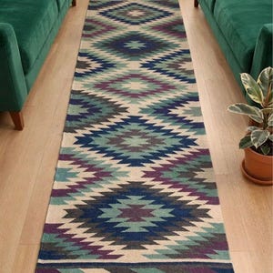 May include: A long rectangular runner rug featuring a repeating geometric diamond pattern in shades of teal, dark blue, purple, grey, and cream. The rug has a flat-woven texture and is finished with short, light brown fringe tassels on both ends.