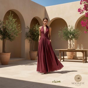 May include: A floor-length, deep burgundy sleeveless dress featuring a plunging V-neckline, a fitted bodice with a gathered waist, and a full, flowing skirt with side pockets. The fabric has a subtle, textured appearance.