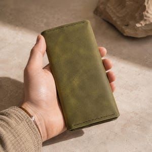 Op de afbeelding: Een rechthoekige olijfgroene lederen portemonnee met een gladde, matte afwerking en zichtbare donkere stiksels langs de boven- en onderrand. De portemonnee heeft een slanke, verticale vormgeving en een egale, effen kleur over het gehele oppervlak.