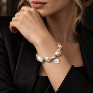 Puede incluir: Una pulsera con perlas barrocas blancas irregulares intercaladas con pequeñas cuentas verdes y detalles en tono dorado. Un dije redondo de metal plateado cuelga de la pulsera, grabado claramente con el nombre de la marca VelaPearl en negro.
