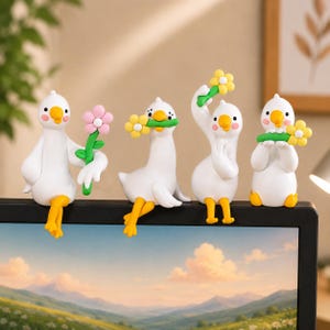 May include: A set of four white goose figurines with orange beaks and feet, each featuring pink cheek accents. The geese are posed holding colorful flowers, including pink and yellow blossoms with green stems, designed to perch on a monitor edge.