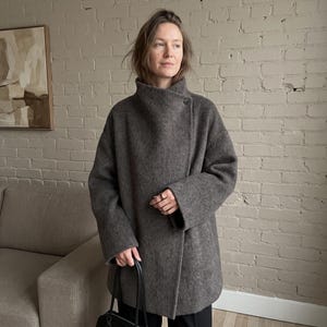 Puede incluir: Un abrigo de mezcla de lana gris carbón con cuello alto, un solo botón visible en el cuello, mangas largas y una silueta recta y relajada que llega hasta la mitad del muslo, usado sobre pantalones oscuros.