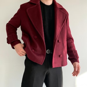 Könnte beinhalten: Eine tiefburgunderrote zweireihige Jacke mit breitem Revers, langen Ärmeln mit geknöpften Manschetten und passenden stoffbezogenen Knöpfen. Die Jacke wird über einem schwarzen Rippenpullover und einer schwarzen Hose mit schwarzem Gürtel mit Silberschnalle getragen.