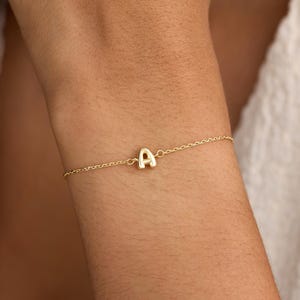 Könnte beinhalten: Ein zartes Goldkettenarmband mit einem einzelnen, mittig platzierten Buchstaben-Anhänger A aus poliertem Gold. Das Armband besteht aus einer feinen Gliederkette, die sich um das Handgelenk legt und ein schlichtes Design mit metallischem Glanz zeigt.